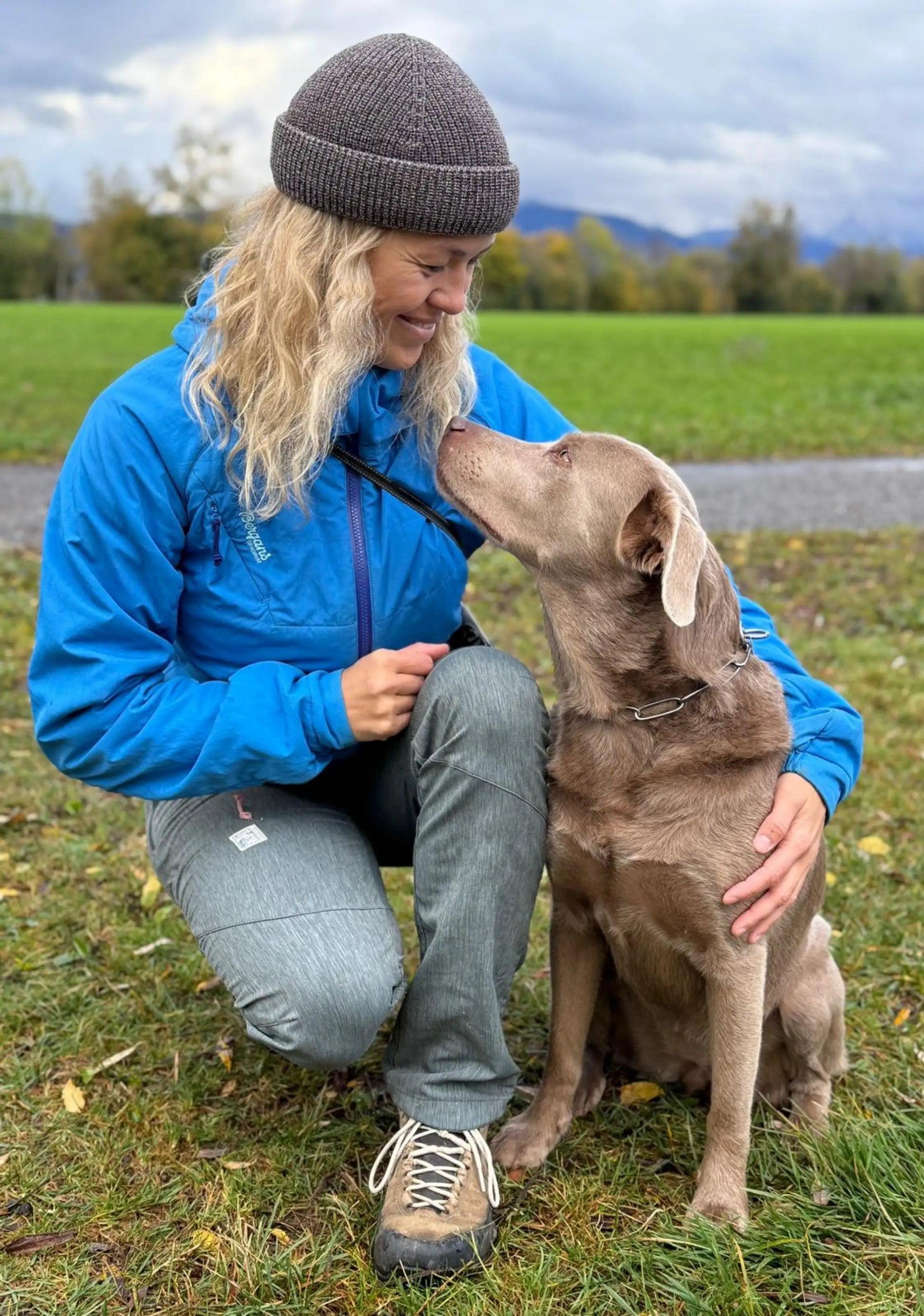 Frau mit langen blonden Haaren und Silbernen Hund schauen sich an. Sie trägt die Fisherman Beanie von ArmyBug in Braun/Weiss meliertem Garn.