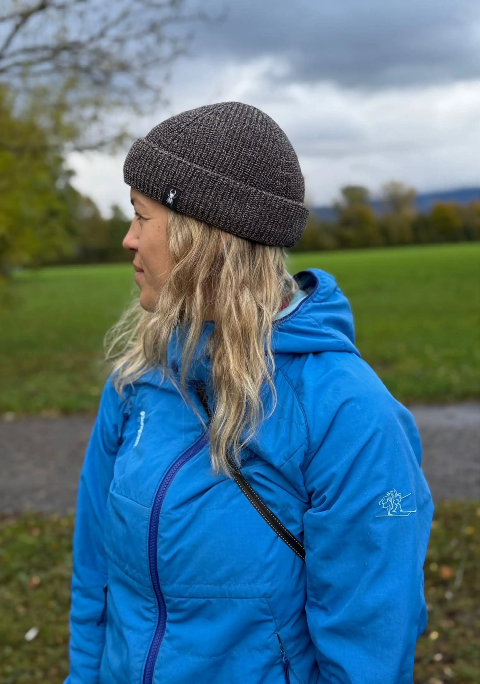 Frau mit langen blonden Haaren und blauer Jacke trägt Braun mellierte Fisherman Beanie von ArmyBug, man sieht den ganzen Oberkörper von der Seite.